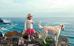 girl in white jacket near brown dog on cliff near body of water during daytime