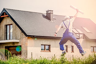 man wearing blue overalls with paper roll jumping near white concrete house at daytime HD wallpaper