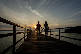 silhouette of man and woman holding hands walking on brown wooden dock during golden hour