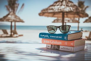 black framed wayfarer-style sunglasses on top of three stack books on beach in selective focus photography
