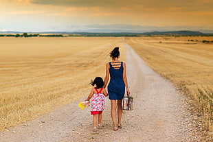 woman in blue dress holding a girl in pink dress walking on dirt road