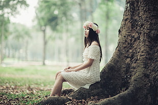 sitting woman wearing floral boho headband and white tiered off-shoulder dress behind tall tree