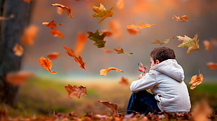 selective focus photo of boy wearing white hoodie sitting on ground with maple leaves