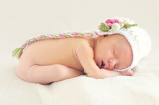naked baby in white,green and pink knitted  cap lying on white textile