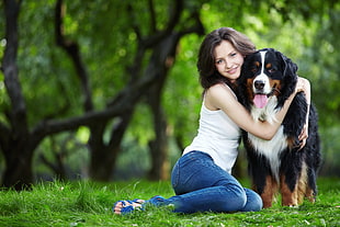 photo of woman in white sleeveless top and blue jeans hugging black, brown, and white long coat dog