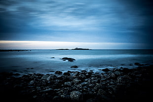 calm sea with stones on seashore under cloudy sky HD wallpaper
