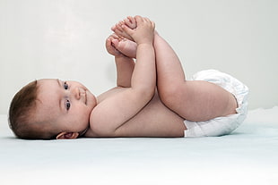 baby in white diaper lying on white textile holding feet