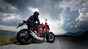 man wearing black leather jacket and black full-face helmet leaning on red and black sports bike under cloudy sky
