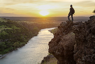 man wearing backpack standing on rock cliff facing sunset infront of river, sacramento river