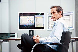 man in white dress shirt sitting near computer monitor while holding a pen