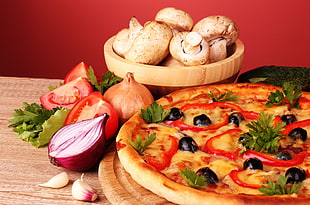red bell pepper and black olives toppings on pizza near brown wooden bowl with mushrooms on brown wooden table