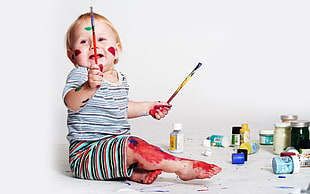 photo of baby holding two paint brushes sitting near paint bottles