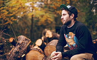 man wearing black graphic printed crewneck and cap sitting near wood logs during daytime