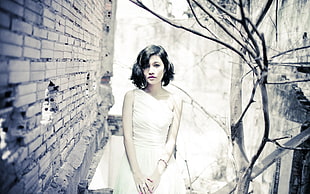 grayscale photography of woman in white one-shoulder sleeveless dress beside bare tree and brick wall