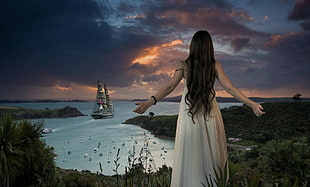 photo of woman in white sleeveless dress facing sea with sailing ship
