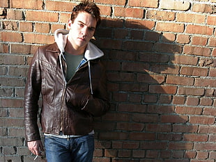 man in brown leather jacket leaning on brick wall