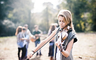 woman wearing grey sleeveless coat plays a violin with people at background selective focus photo