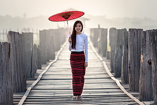 woman in white long-sleeved shirt and red striped skirt holding umbrella walking on gray wooden pathway