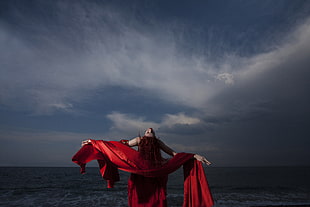 woman in red dress standing near body of water under gray sky