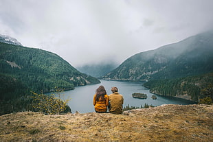 two person sitting on cliff near lake surrounded by mountains, photography, nature, landscape, couple