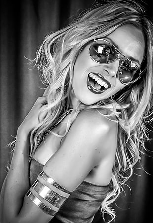 grayscale photography of woman standing while smiling with aviator style sunglasses