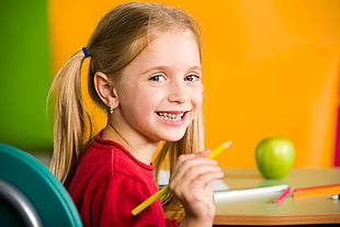 girl in red crew-neck shirt holding pencil fronting green apple