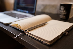 selective focus photography of brown notebook and gray click pen on top of brown wooden table