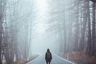 woman in black jacket standing in the middle of concrete road surrounded by tall bare trees covered with fog