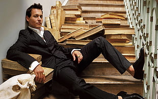 man wearing black blazer, white dress shirt, black dress pants, with black dress shoes sitting on stairs leaning on wall surrounded by brown hardbound book