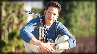 sitting man wearing blue sweater and white denim jeans wearing watch with silver saxophone during daytime