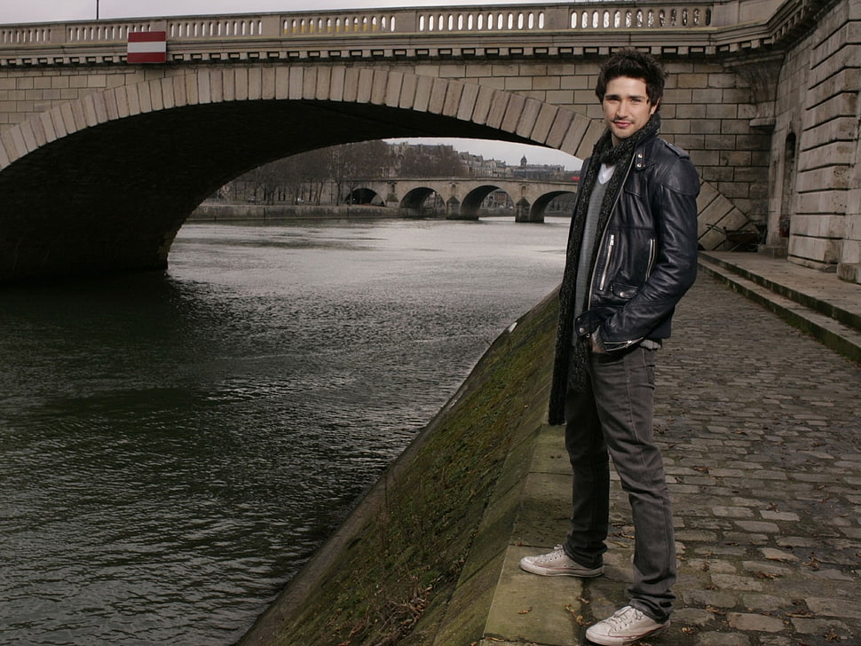 man in black leather zip-up jacket standing near concrete bridge during daytime HD wallpaper
