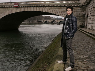man in black leather zip-up jacket standing near concrete bridge during daytime