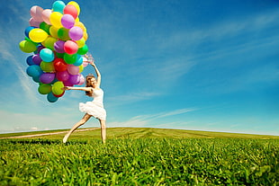 woman jumps on green grass holds assorted-color balloons under blue sky at daytime