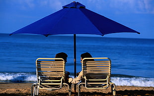 couple sitting on white steel folding bed near in the beach under blue umbrella during sunset