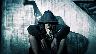 man in black hoodie sitting beside wooden wall