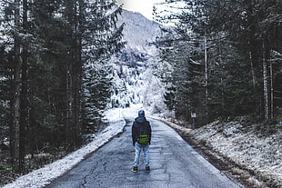 person in black jacket with black backpack standing on grey concrete road near trees with snow during daytime