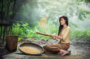 woman sitting on land holding brown wicker tray with grains