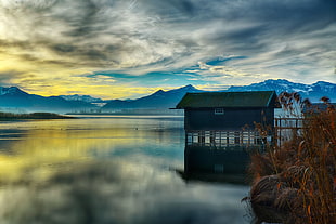 grey and green wooden house on center field water with mountain hills under cloudy sky during daytime HD wallpaper