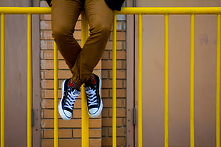 person with brown pants and black-and-white lace-up low-top sneakers sitting on yellow metal railings