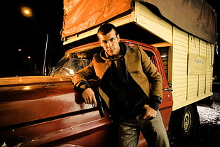 man in brown jacket, black shirt, and gray denim bottoms leaning on red and white box truck