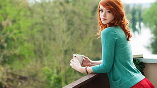 selective focus photography of woman wearing green cardigan leaning on wooden rail while holding white mug