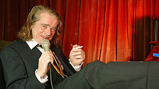man in suit jacket and dress pants holding microphone and cigarette while sitting