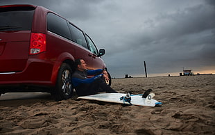 man sitting on surfboard leaning on red vehicle parked on shore