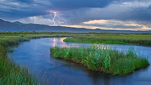 river between green grass field with a view of a lightning and mountains afar at daytime