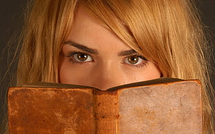 portrait photo of woman with blonde hair covering her face with brown hardbound book