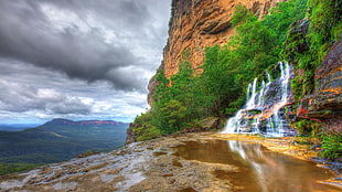 small waterfall under mountain and tress during daytime