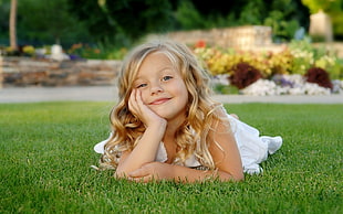 blonde-haired girl in white dress laying down on green grass with head on right hand smiling