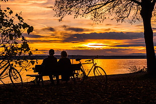 silhouette of two person sitting on bench beside bicycle by the body of water