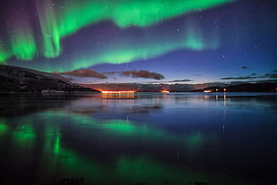 green aurora on sky reflected on body of water in distant of structures with lights