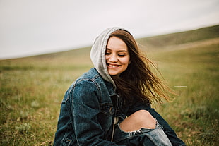 photography of brown haired woman wearing blue denim jacket seating on green grass field during time